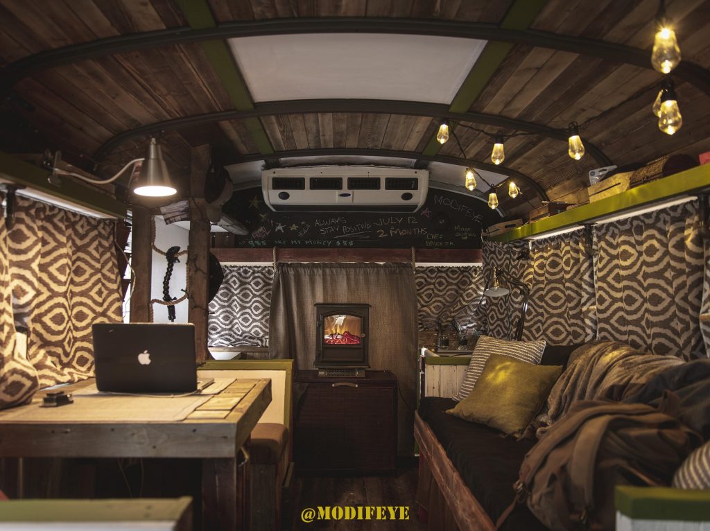 Cozy interior of a skoolie with a fireplaces, curtains drawn, and a desk with a Macbook. 